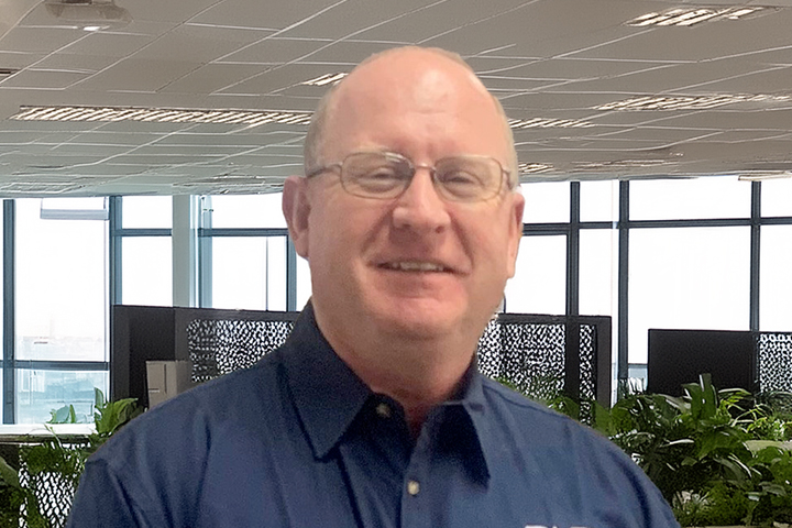 The image shows Stuart McKinder in a dark blue TAFE NSW button-up shirt. He is wearing glasses and smiling at the camera. He is in a warehouse setting.