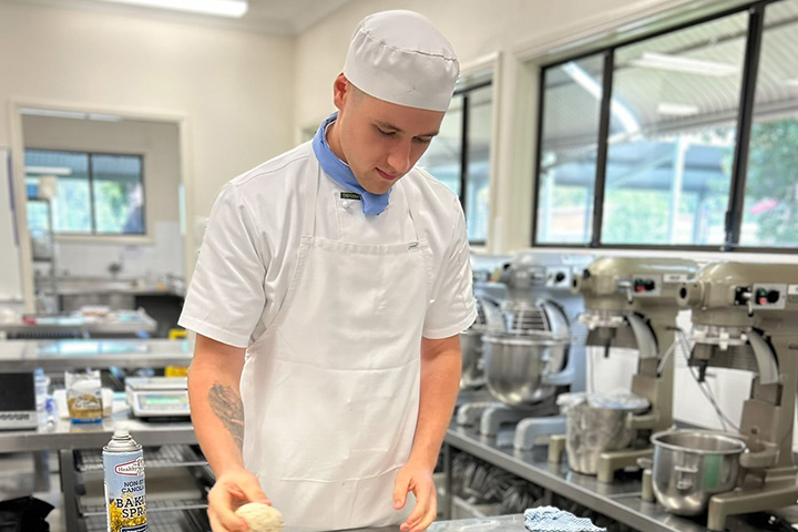 Connor Beisler is wearing a white uniform, white apron, bakers hat and blue neck tie, while working with dough in the TAFE NSW commercial bakery. 