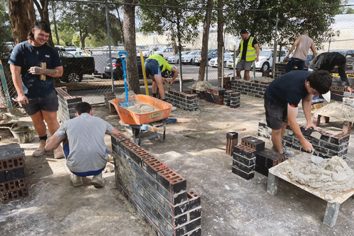 A group of people laying bricks and mortar outdoors, constructing low training walls on a concrete pad with tools and a wheelbarrow.