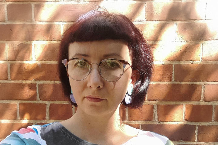 The TAFE NSW Visual Art and Design teacher stands against a sun-dappled wall with a closed-mouth smile. She wears glasses, creative earrings, and a colorful t-shirt.