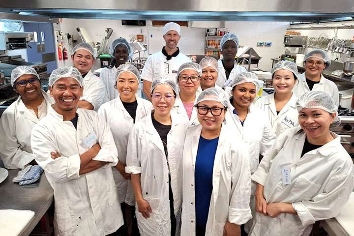 A group of AMEP students and their teacher wearing white kitchen uniforms and hairnets smile together in a commercial kitchen.