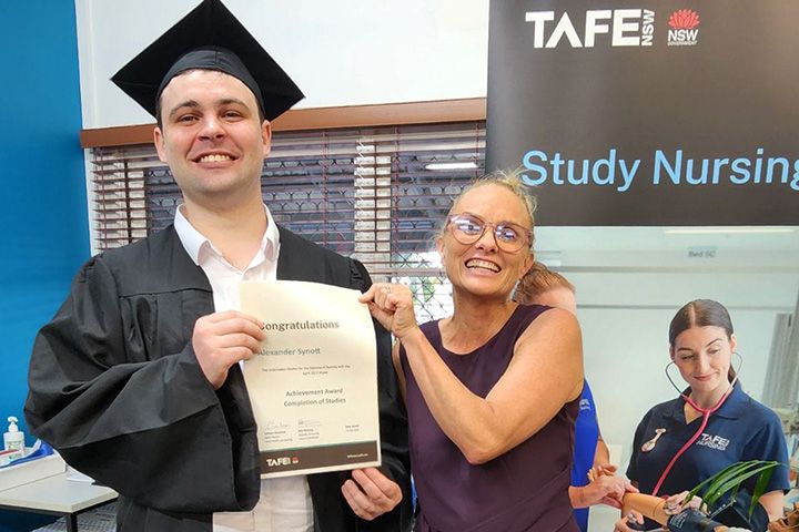 Alex Synott, a young man wearing a black graduation cap and gown, smiles while holding a certificate. Beside him, a woman with glasses and tied-back blonde hair proudly holds his arm and smiles. TAFE NSW banner in the background reads "Study Nursing."