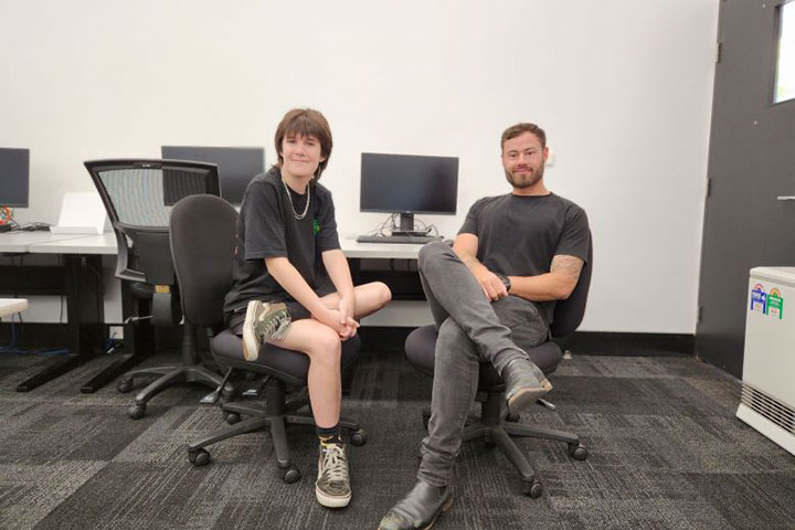 Two students sitting on computer chairs in a computer lab, smiling at the camera.