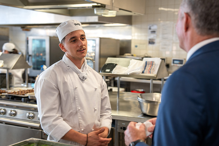 A TAFE NSW Commercial Cookery student stands in a kitchen facility. He is wearing a white chef’s uniform and hat, and is engaged in conversation with a man in a blue jacket.