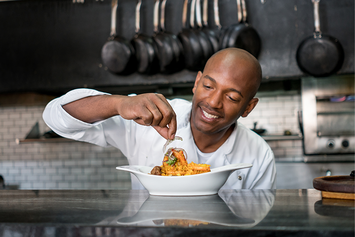 Chef sprinkles garnish on a plate of food. He is smiling and in a commercial kitchen.