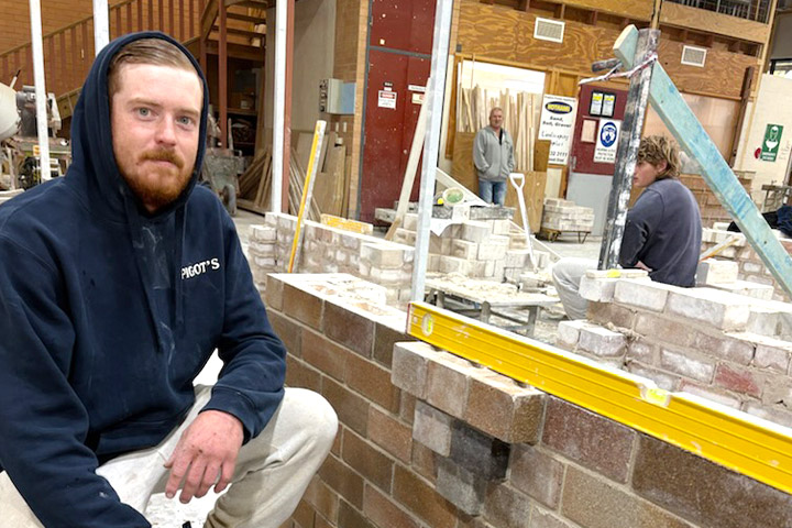 27 year old Cody squats beside a low brick wall, holding a tool. He wears a dark blue hoodie and white tracksuit-pants and smiles with a closed mouth, looking at the camera.