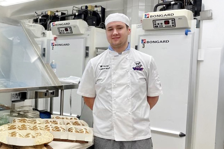 Connor Beisler in a white baker’s uniform, smiling in a bakery with hot cross buns.