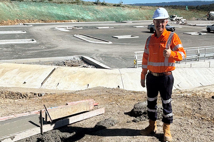Former Central Coast furniture salesman Jaydon Brown, wearing an orange  high-vis shirt, blue work pants, boots and hardhat, standing on a raised bank of earth in front of a major highway construction project and giving a “thumbs up”. 