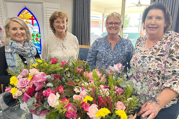 Four women smile while standing behind a large arrangement of colourful flowers, including roses, daisies, and native blooms. They are indoors, with a stained glass window and curtains in the background.
