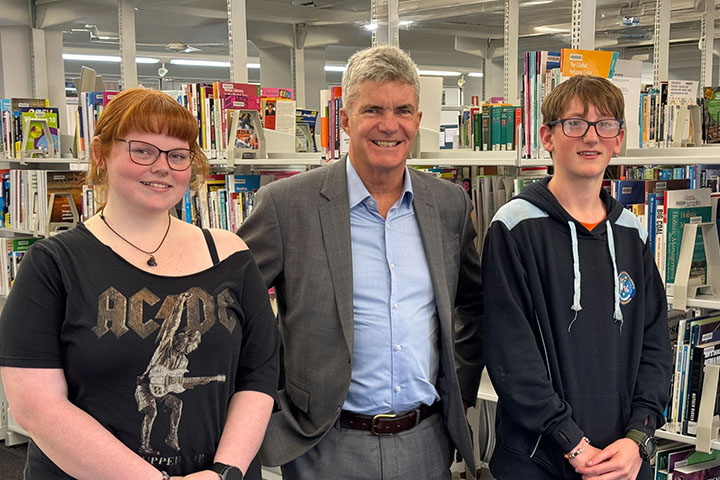 The Minister for Skills, TAFE and Tertiary Education, Steve Whan (centre), stands smiling between students Alice Hale (left) and Nathaniel Davis (right) at TAFE NSW Goulburn. They are in a library setting, with bookshelves full of educational materials in the background.