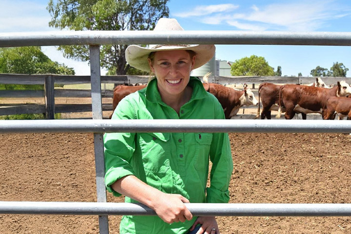 Farmer and TAFE NSW butchery student Hayley Patterson stands leaning on a stockyard fence with a small herd of cattle behind her.