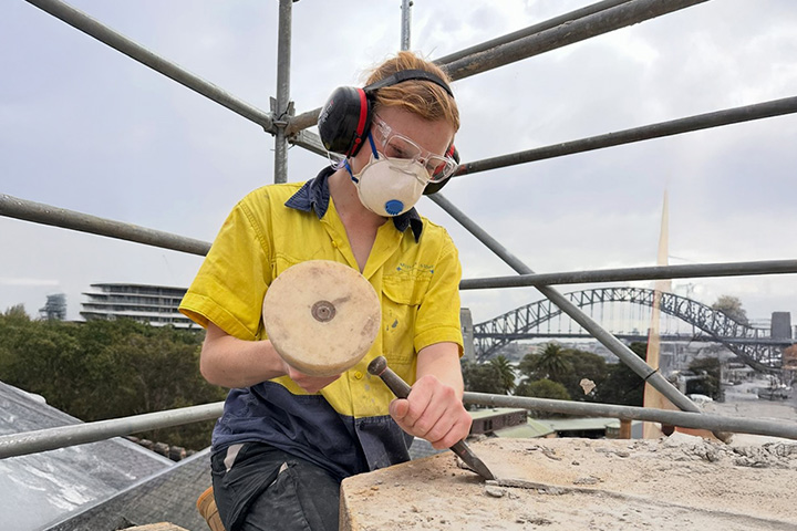 Haley Sharp undertaking stonemasonry conservation work at Government House. She is up high, holding tools and wearing protective equipment with scaffolding behind her and the Sydney Opera House further in the background.