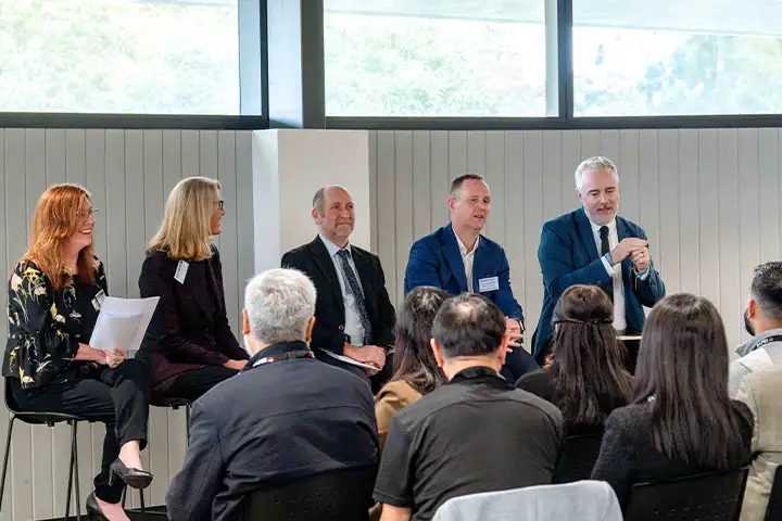 A panel of industry speakers made up of two women and three men sit in front of an engaged audience. Behind them is a white panelled wall and high-level windows with light streaming through.