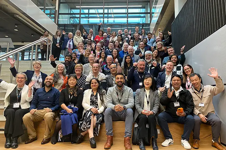 Industry Accelerator program attendees smile and wave for a photo at the launch event at TAFE NSW Meadowbank. They sit in a light filled auditorium style space on wooden stairs.