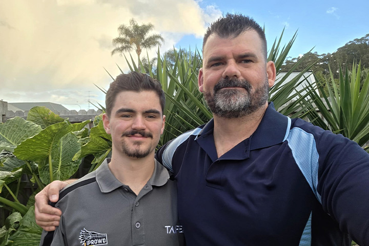 TAFE NSW IPROWD graduate Jackson Cooper and his father Aaron, with his arm around Jackson’s shoulders, stand smiling at camera in front of green plants and a cloudy blue sky.