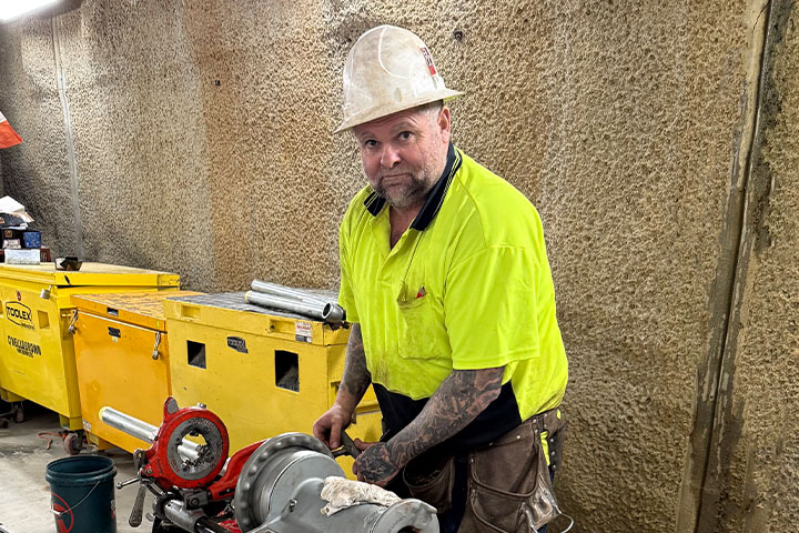 Jason Day smiles at the camera while working indoors surrounded by tools and equipment. He wears a hi-vis, yellow, polo shirt, a hard-hat and a tool belt.