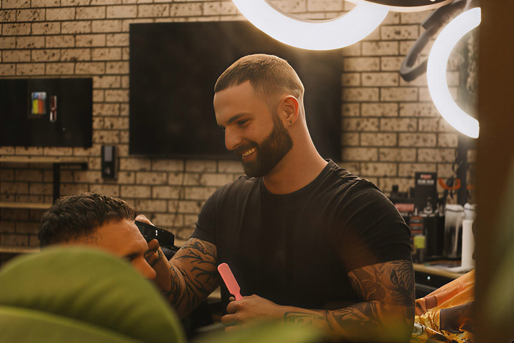 TAFE NSW graduate Joshua Landi gives his client a haircut. He is wearing a black t-shirt and smiling, with clippers in one hand and a brush in the other.