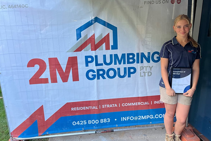 Young blonde woman in a 2M Plumbing Group uniform stands next to a company banner, holding a certificate that reads "Completion of 100 days.