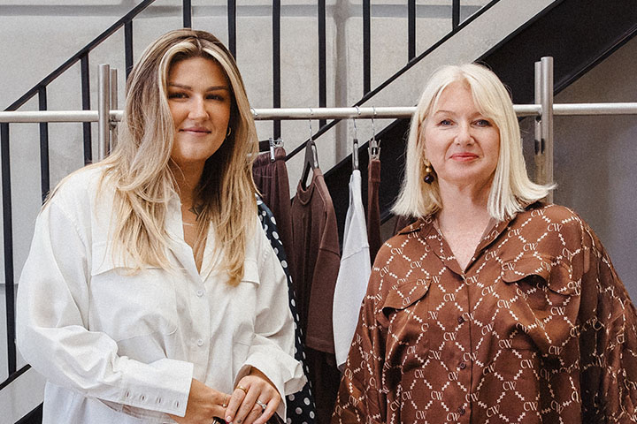 Kayla Reynolds and Dyan Thais stand side by side subtly smiling at the camera. They stand in front of a retail clothing rack.