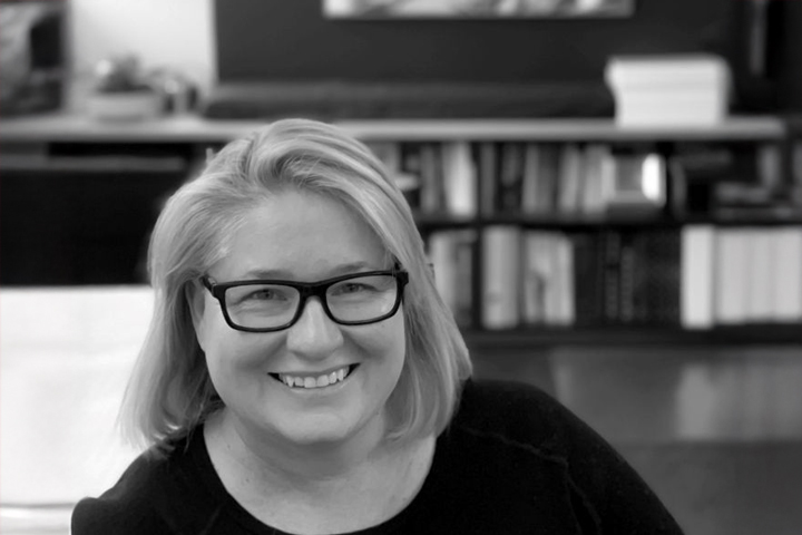 A smiling woman with shoulder-length blonde hair and black-framed glasses sits in front of a bookshelf and an abstract painting; the image is in black and white.