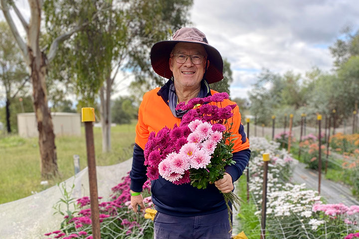 Laurence Tockuss stands between flower beds. He wears work clothes, holds a bunch of bright pink and crimson flowers, and smiles at the camera.