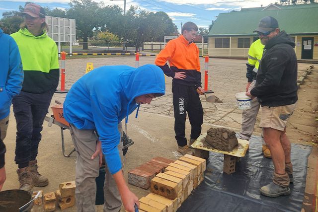 Teenage boy in blue jumper and hood stacking the bricks. Five other boys in jumper and pants watching and assisting the boy with bricklaying. 