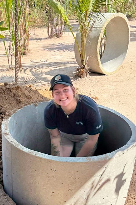 Maddie Tuchin kneels inside a concrete cylinder while smiling. The background features sand and palm trees.