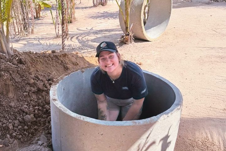 Maddie Tuchin smiling and kneeling in a large pipe. The background features sand and palm trees.