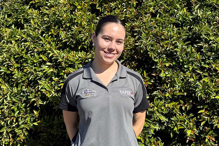 Makayla Harte, a proud Ngunnawal woman, stands smiling at the camera in front of a large bush with green leaves. She wears a grey polo shirt with TAFE NSW and IPROWD logos.