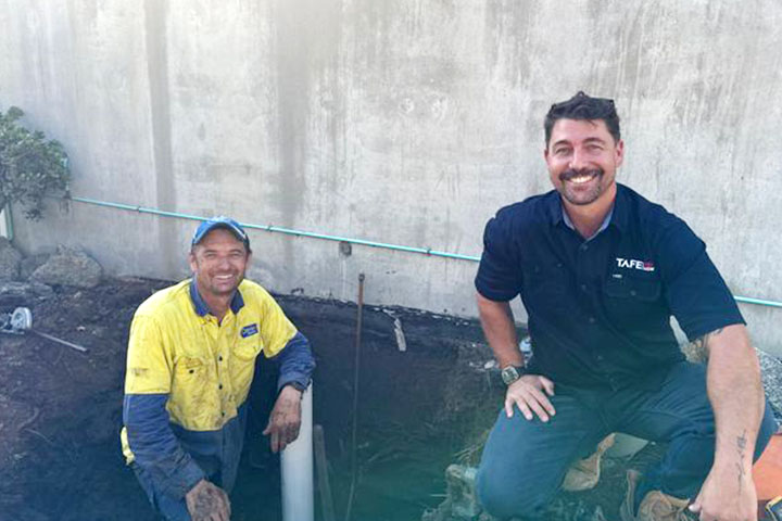 Two men kneeing in the dirt beside a white pipe, smiling at the camera.