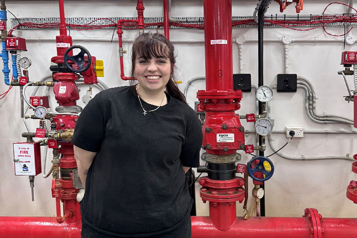 TAFE NSW student Montanna Weir wears black jeans and a black t-shirt and stands in front of a large, red fire sprinkler system, smiling and looking at the camera.