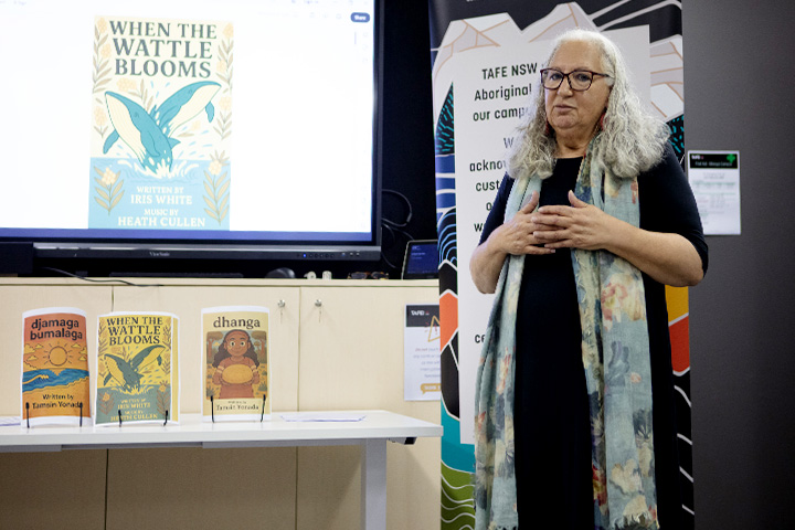 Aunty Iris White at the Dhurga language resources launch, with books on display and a screen showing the book ‘Where the Wattle Blooms'.
