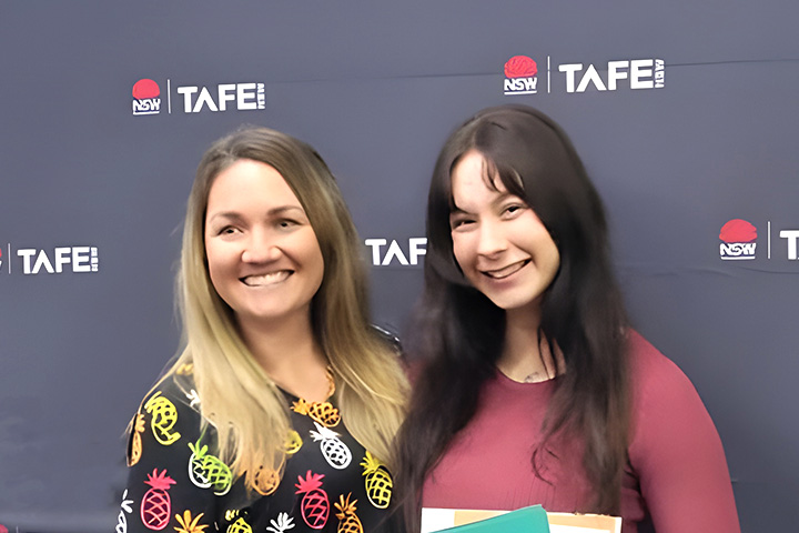 TAFE NSW teacher Hayley Strath and graduate Natalie Lucas stand together, smiling, in front of a black TAFE NSW backdrop. 
