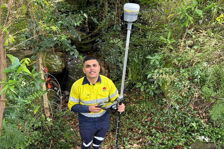 Nick Baran wears a high-visibility yellow and navy work uniform with reflective stripes. He is standing in a lush, wooded area holding surveying equipment.