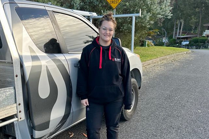 Nicole Howarth stands next to a work ute with a ladder on top, wearing a black hoodie and work boots. Trees and houses line the quiet street behind her.