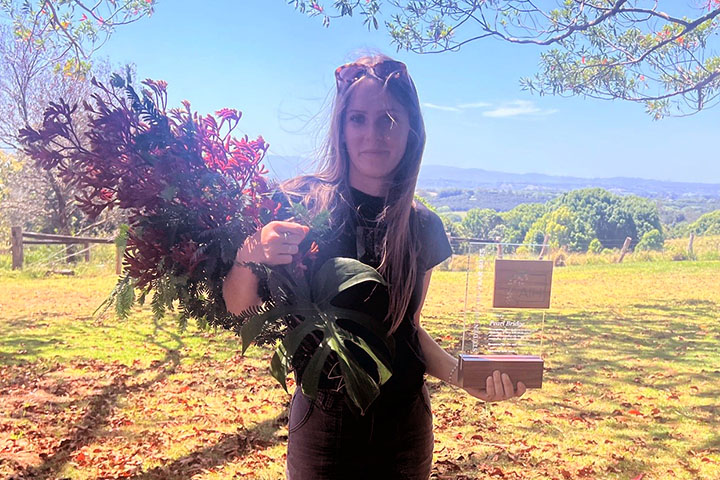 Pearl Bridge, wearing jeans and a t-shirt, stands under a tree in a rural setting. She’s smiling at the camera holding a large bouquet of flowers and her award.