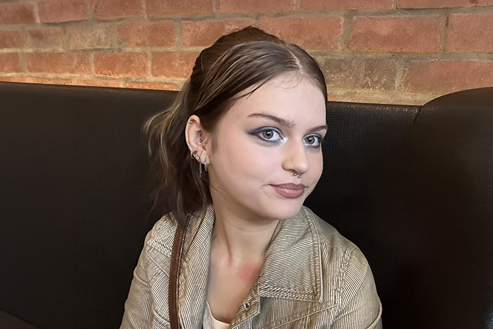 Quinn Street sits on a sofa, head turned to the right. She has a light smile and appears to be looking at someone behind the camera. She wears a striped, light brown jacket and has her brown hair tied back in a knot.