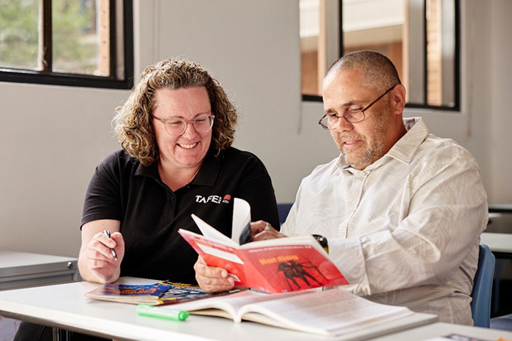 TAFE NSW teacher sits beside an adult learner at a desk, smiling while supporting him to read a book and review learning materials together in a bright classroom, highlighting adult literacy support through the Reading Writing Hotline.
