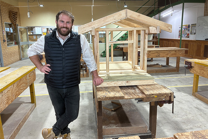 Sam Horn, Carpentry and Construction Teacher is wearing black pants, white shirt and black vest with work boots. He is smiling while learning against a work bench in the carpentry room. A small house-like structure sits behind him on the bench.