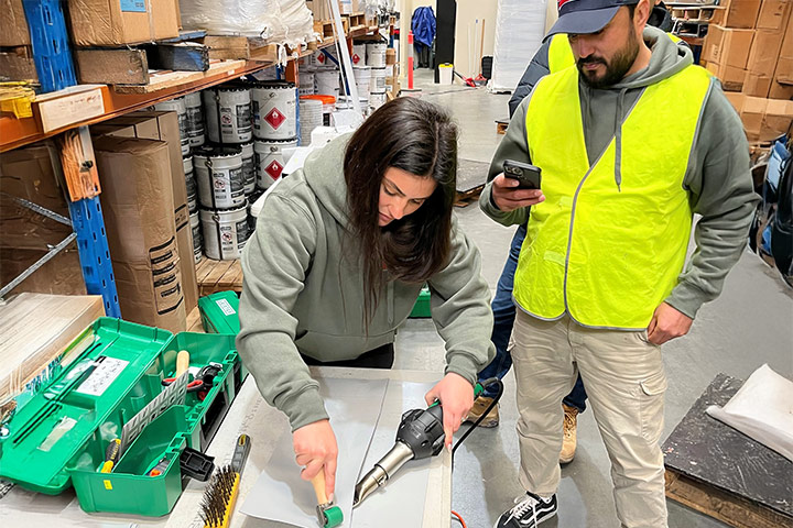 Scheriya stands at a work bench using waterproofing tools including a roller and a hot air gun, watched by a tradesman in a high-vis vest.