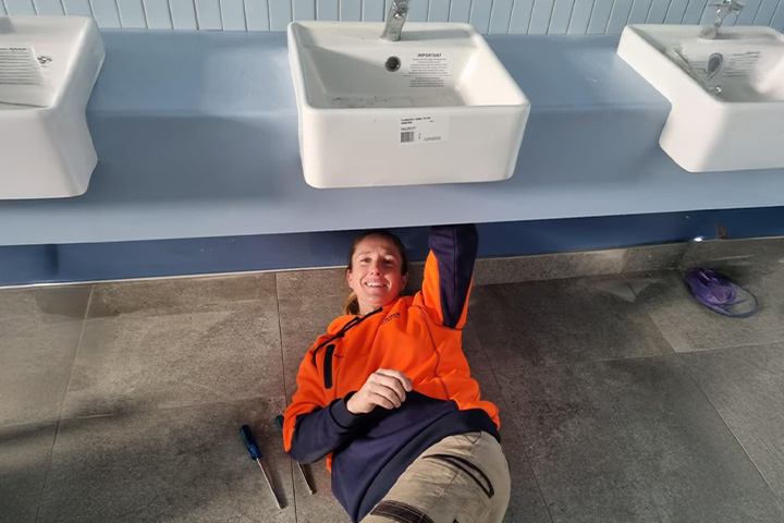 Shelley Knight is laying underneath a row of bathroom sinks looking towards the camera smiling. She is wearing an orange and navy high-vis jumper and cream cargo pants. Her hair is tied back
