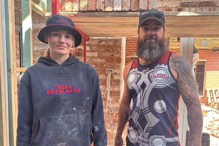 Sophie Carver standing next to her dad/boss Wes Carver at a job site. Sophie wears a grey bucket hat and grey hoodie that reads ‘Thor’s Bricklaying’ and her dad wears a singlet top with the same branding.