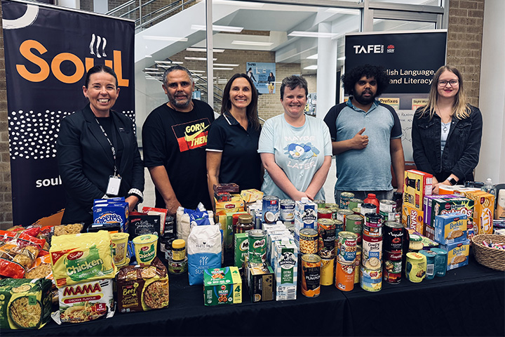 Six people stand smiling behind a table filled with non-perishable food donations at TAFE NSW Newcastle, supporting a charity drive for Soul Hub.