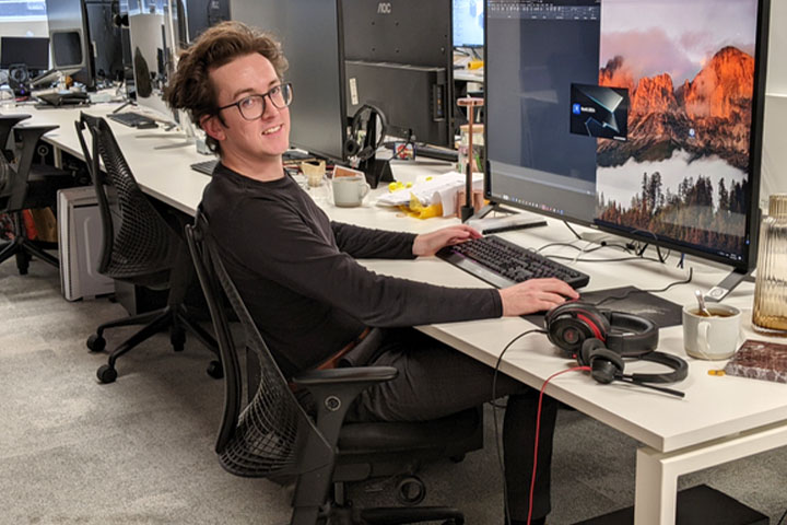 A student sits at a computer desk, wearing glasses, smiling with his hands on his mouse and keyboard.