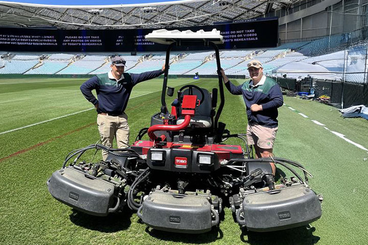 Students Luke Urkanci and Wade Buhler lean casually against a turf management tractor they’re using to prepare the Accor Stadium for the NRL ahead of the Grand Final. It’s a blue sky, sunny day. They are both smiling at the camera under sun caps and wear beige pants, and blue and green long-sleeved polo shirts. Sunglasses are perched on their visors.