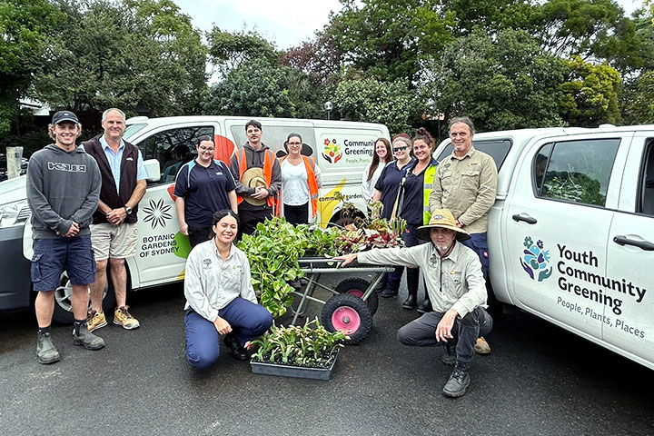 TAFE NSW Padstow students pose together in front of two Community Greening vans with a wheelbarrow full of plants.