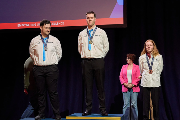 Three young adults stand on a winners' podium, wearing medals around their necks at an awards ceremony. The gold and silver medallists wear cream button up shirts with a TAFE NSW logo and black pants. The backdrop includes a red banner with the words "Empowering Skills Excellence."