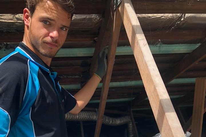 Tom is wearing a navy blue t-shirt with sections of light blue. He is looking at the camera with one gloved hand resting against a roof truss.