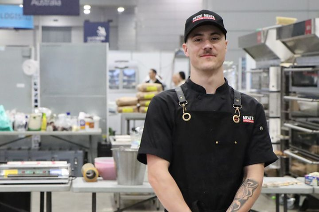 Tyler Walters standing, facing toward the camera with a butchery behind him. He wears a black cap, black t-shirt, and a black apron on top.