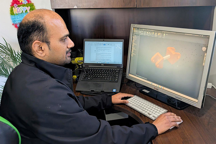 A focused student working at a desk on a computer. A large monitor shows an AI generated image, while a smaller laptop on his left-hand side displays a text document.